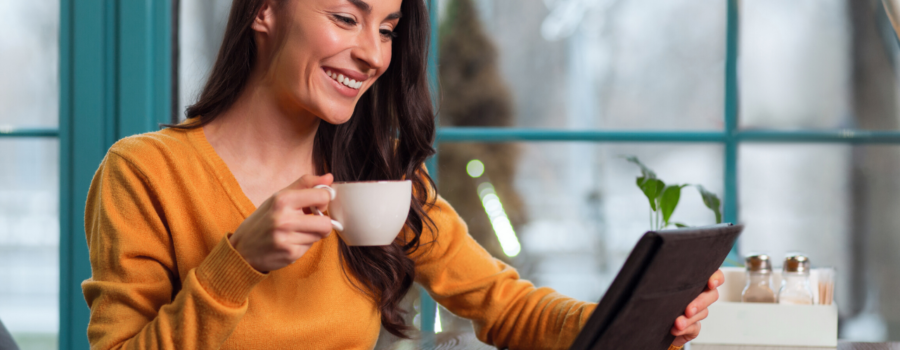 A woman looking at her tablet, smiling, holding a cup of coffee