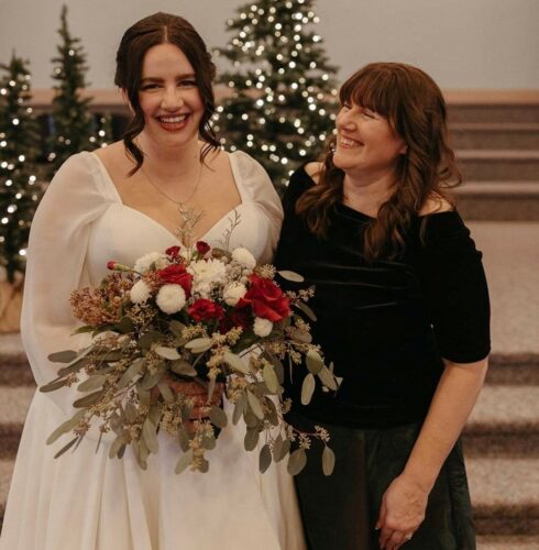my daugher and I, she is wearing a wedding gown and holding flowers, I am looking at her smiling, there are holiday lit trees behind us. Credit for the photo to Roxanne Duke Photography.
