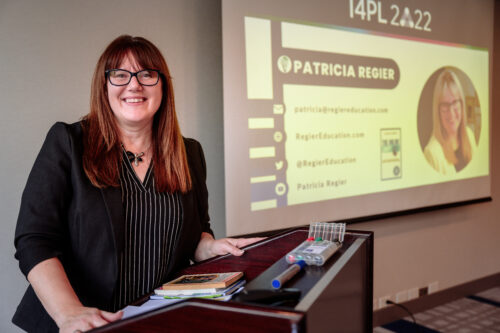 Patricia Regier Brown Hair, Necklace, Black and White Stripped Shirt and Black jacket, Wearing glasses and smiling. PowerPoint slide in background and standing at a podium.