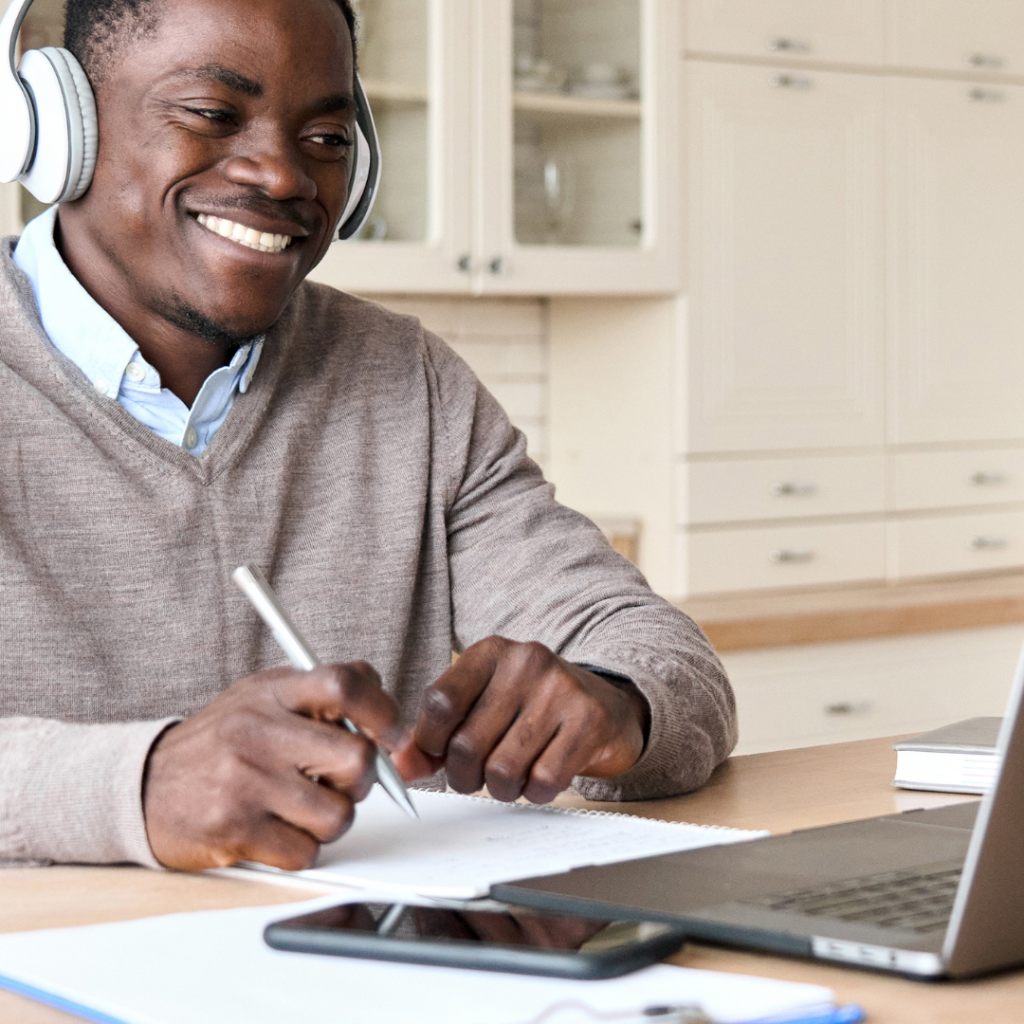 Black man with a headset on, looking at a laptop, phone on seide, pen in hand, attending a Zoom meeting. Smiling
