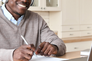 Black man with a headset on, looking at a laptop, phone on seide, pen in hand, attending a Zoom meeting. Smiling