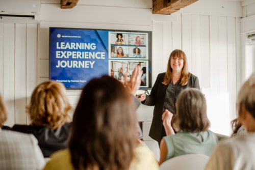 Patricia speaking to a group. She is a white woman with brown hair wearing a black and white striped top and black pants. PowerPoint slide is on the TV on the wall.