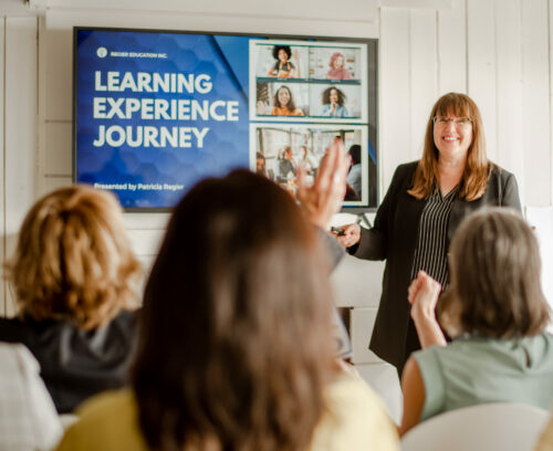 Patricia speaking to a group. She is a white woman with brown hair wearing a black and white striped top and black pants. PowerPoint slide is on the TV on the wall.