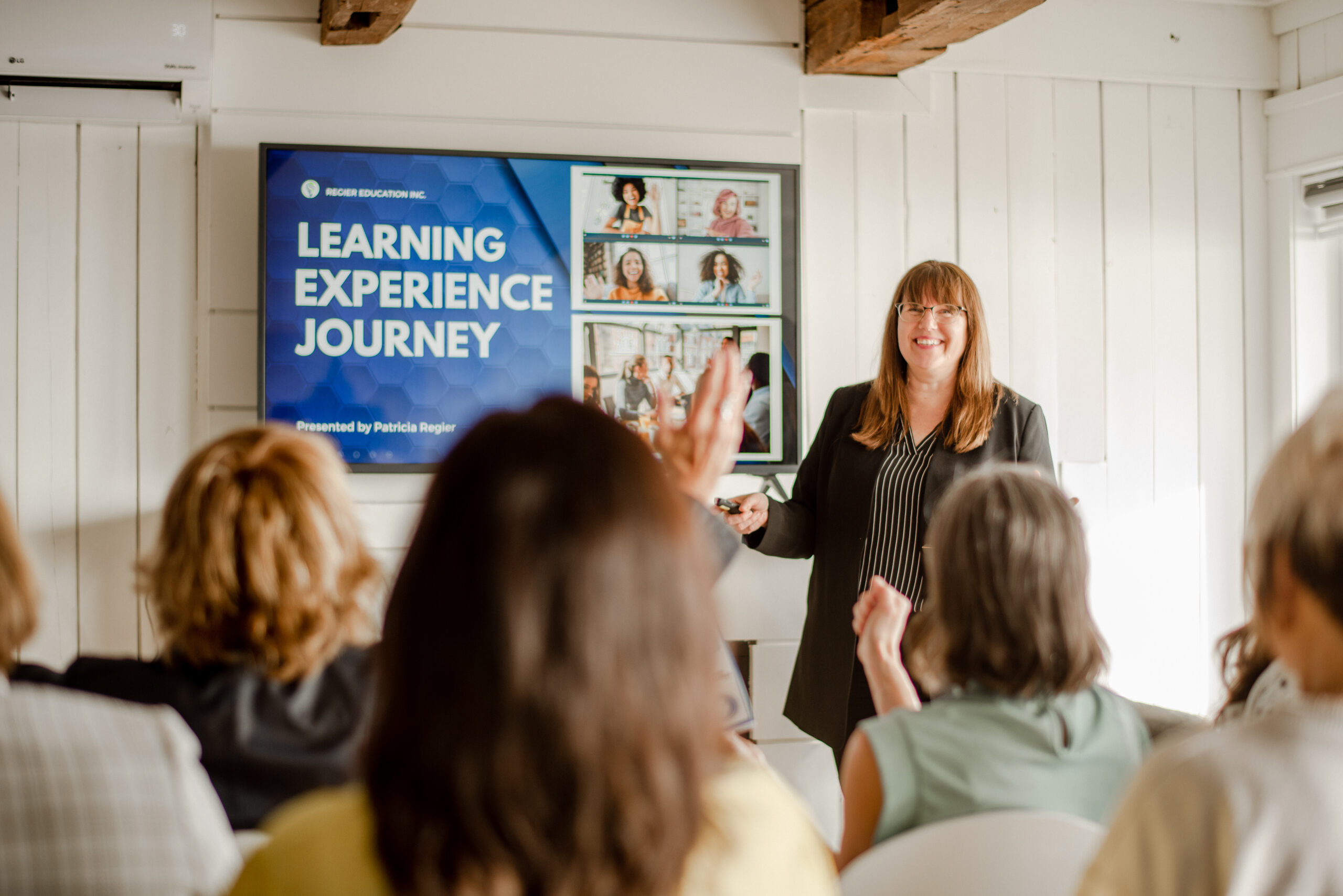Patricia speaking to a group. She is a white woman with brown hair wearing a black and white striped top and black pants. PowerPoint slide is on the TV on the wall.