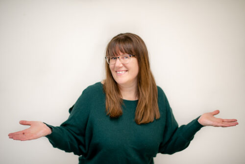 Patricia Regier, a white woman with brown long hair and glasses. Smiling, and wearing a green sweater. Arms are wide and hands open facing upwards.