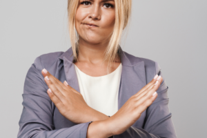 White woman with blond hair, face expression mouth to the side, and arms in an X formation with hands to demonstrate No. Person wearing grey suite jacket and white top. No to WebEx Expression.