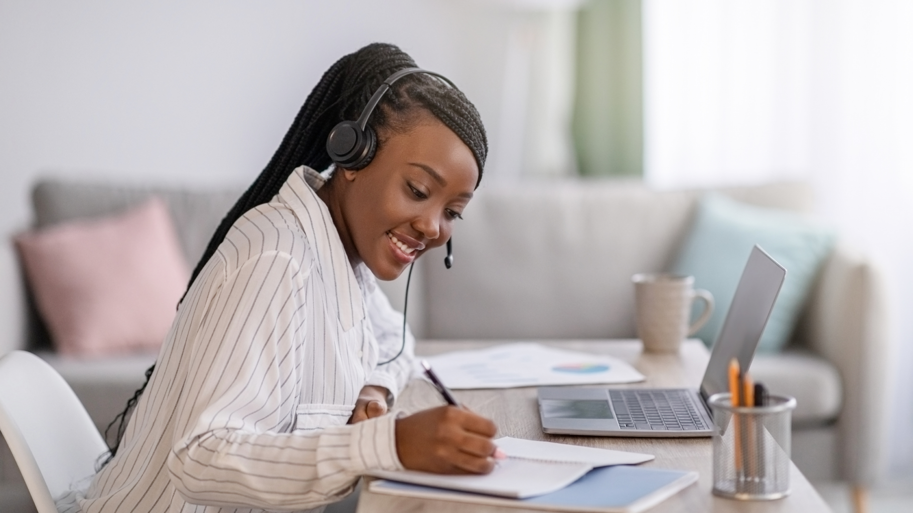 black woman taking an online course, smiling, writing in a note book, head phones on, and laptop open.