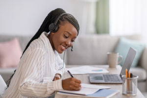 black woman taking an online course, smiling, writing in a note book, head phones on, and laptop open.