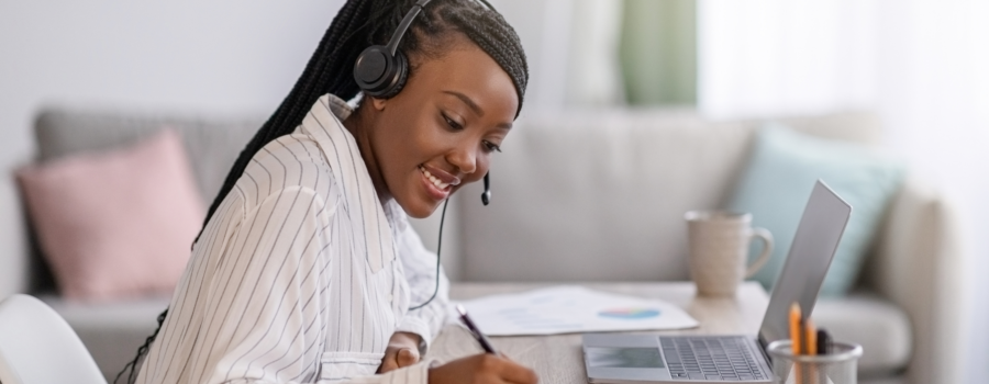 black woman taking an online course, smiling, writing in a note book, head phones on, and laptop open.