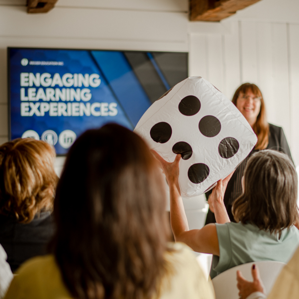 Patricia facilitating a group, words Engaging Learning Experiences is on a tv screen, and a participant is holding a large dice. Celebrate.