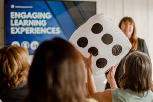 Patricia facilitating a group, words Engaging Learning Experiences is on a tv screen, and a participant is holding a large dice. Celebrate.