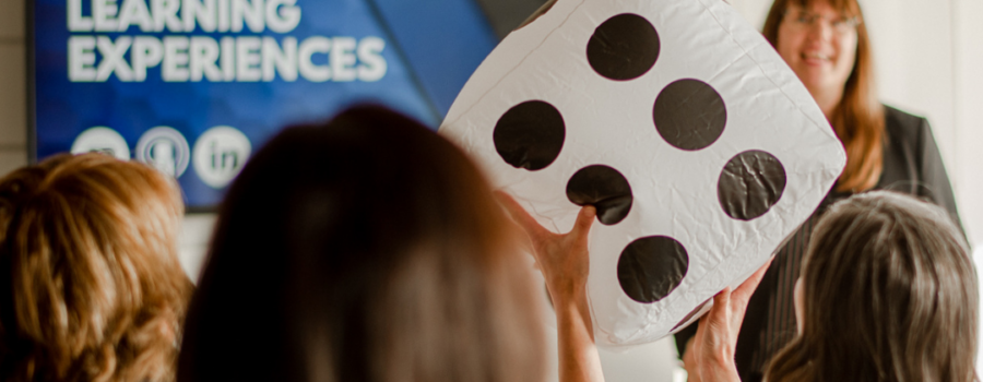 Patricia facilitating a group, words Engaging Learning Experiences is on a tv screen, and a participant is holding a large dice. Celebrate.